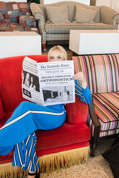 Headshot of Dr. Brooks, orthodontist in Nashville, TN, sitting on a red couch in a blue outfit holding a coming soon newspaper