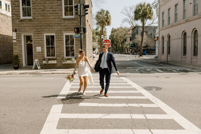 Bride and groom holding hands, walking across a downtown Charleston crosswalk during their stylish city elopement