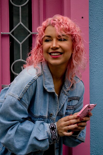 A woman with pink curly hair is holding a phone and smiling.