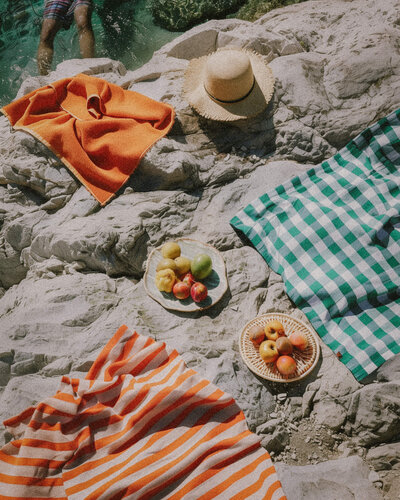 Waterside picnic, with bright orange and white striped towel and Turqouise and white Gingham Towel, plates with fruit and a straw hat
