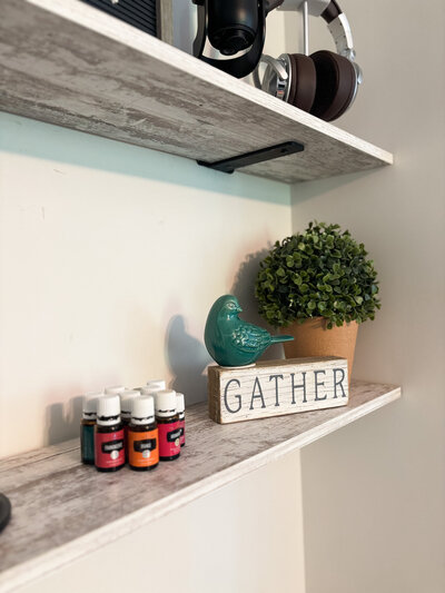 Wooden shelf featuring essential oils, greenery, and a decorative "gather" sign with cozy vibes. 