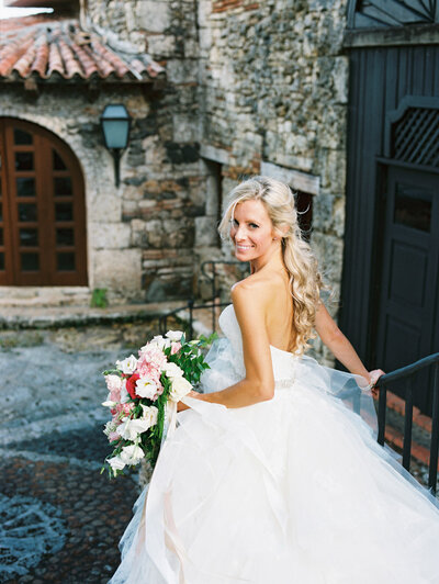 Bride smiling over her shoulder while holding a lush pink and white bouquet in the stone streets of Altos de Chavón — captured by Asia Pimentel Photography.