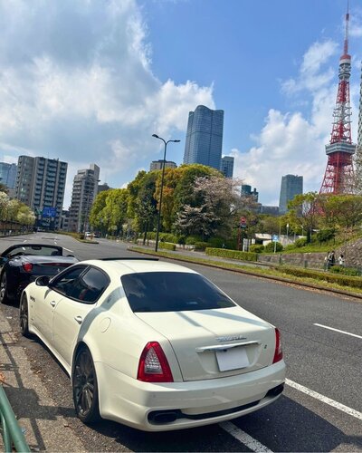 A white Maserati parked on a city street with Tokyo Tower visible in the background under a blue sky with scattered clouds.
