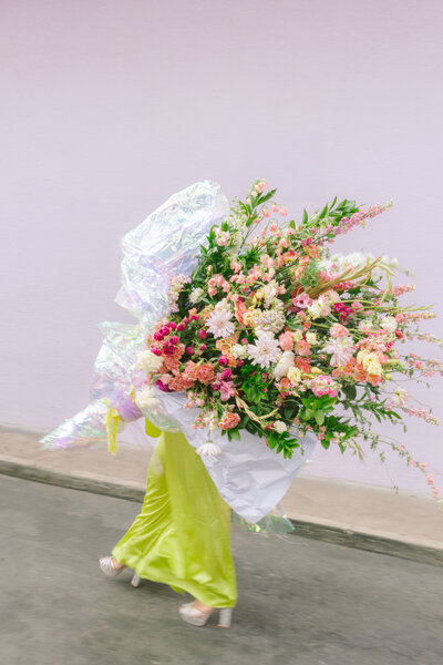 A striking editorial image of a woman in a chartreuse silk dress and glitter heels walking down the street with an enormous, whimsical bouquet of pink and peach flowers wrapped in iridescent foil. Set against a pale lavender wall, the blurred motion adds energy and fashion-forward flair. Captured by Dani Lacey Photographs.