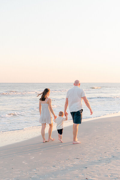 Mom and Dad holding their toddler son's hand while candidly walking on the beach in Hilton Head, SC with Hilton Head Photographer, Lamp and Light Photography.