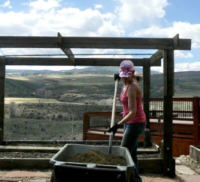 A woman digs into dirt as she works on her landscaping and garden beds with the mountains in view behind her.