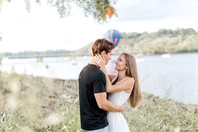 Engaged couple embrace as hot air balloons take off in the background.