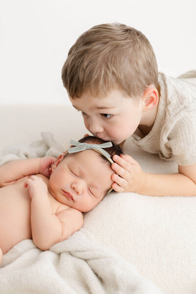 New Mom in beige floral dress with professional hair and makeup done. She is holding her newborn baby up by her face and has her eyes closed. Baby is also sleeping and wrapped in a beige swaddle his feet and hands are showing.