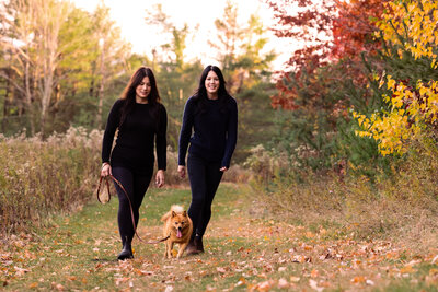 Two women walk their dog  on a trail with fall foliage by  Vermont Family Photographer