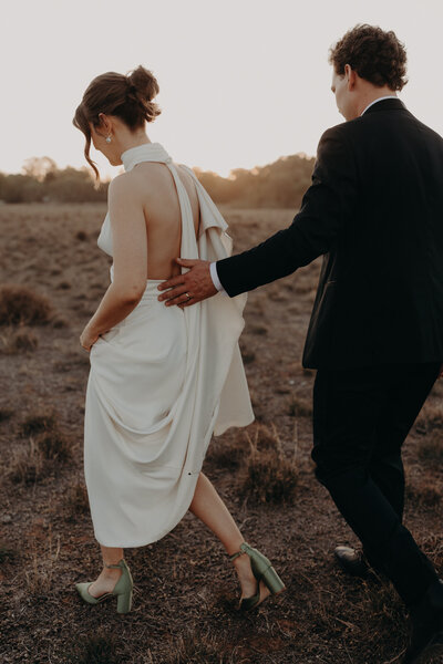 Bride and Groom walk through paddock at sunset. Groom has his hand around Brides waist.