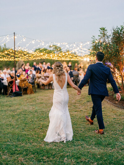 Bride and groom holding hands walking towards all their wedding guests under string lights