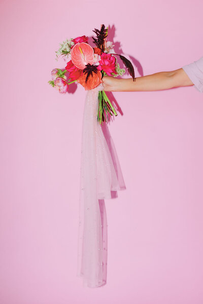 Arm holding a pink bouquet of flowers with white tulle hanging down in front of a pink wall