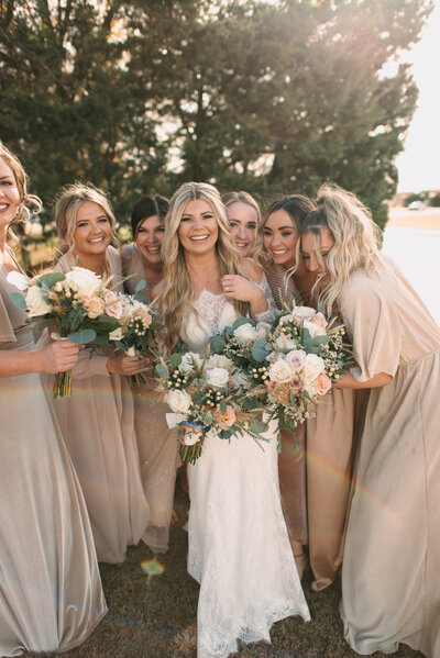 Bride being hugged around by all of her friends while they all hold their beautiful big bouquets and neutral bridesmaids dresses laughing and having fun as they support her on her wedding day