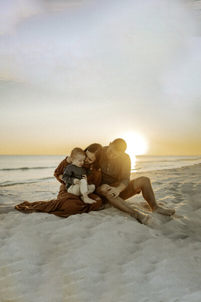 Family Photographer, a man and woman sit closely together on the beach sand at sunset. They admire their toddler son together.