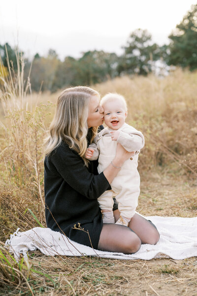 Mom sits on white blanket in brown grassy field kissing smiling baby boy during family photos