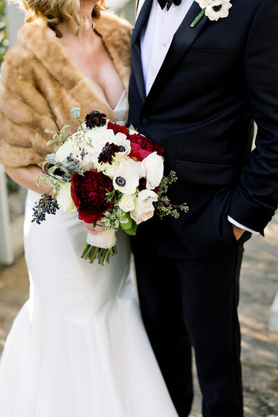 woman in white dress with fur coat holding red and white flowers next to man in black tuxedo
