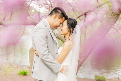 Newlyweds touch foreheads while holding each other under a pink blossoming tree by a lake