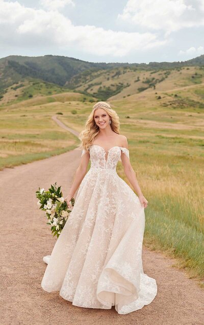 FLORAL-INSPIRED A-LINE WEDDING GOWN A floral-inspired A-line wedding dress, this Martina Liana gown is the definition of a natural beauty! The gown’s structured bodice features a slight plunge, and is adorned with striking floral booms. The neckline extends off-the-shoulder into drapey sleeves that add a bohemian touch to the style. A mix of leaves, vines and floral lace continues throughout the gown, adding to the overall botanical feeling, while the silhouette remains classic. The gown’s sheer bodice features exposed boning and layered French lace, which is continued through the back of the gown. A dramatic train with bold detailing completes the look of the gown effortlessly. This A-line wedding dress is available beaded or unbeaded.