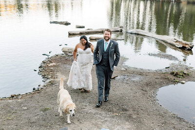 A bride and groom walk along the edge of a lake with their dog while on their mountain wedding day. 