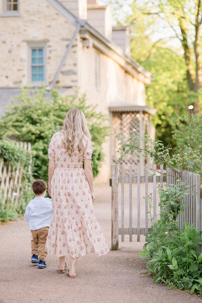 A mother wearing a Doen dress holds her son's hand and walks toward the house at Bartram's Garden.