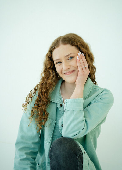 Haylee smiling at the camera with one hand on her knee, touching the side of her face.