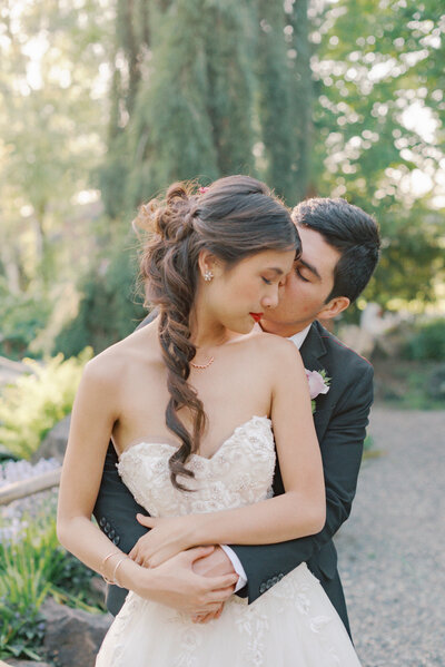 groom with hands wrapped around brides waist giving a kiss from behind