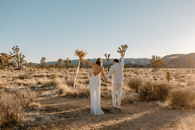 Newlywed couple walking hand in hand through Joshua Tree National Park, raising their arms in celebration during their sunset elopement.