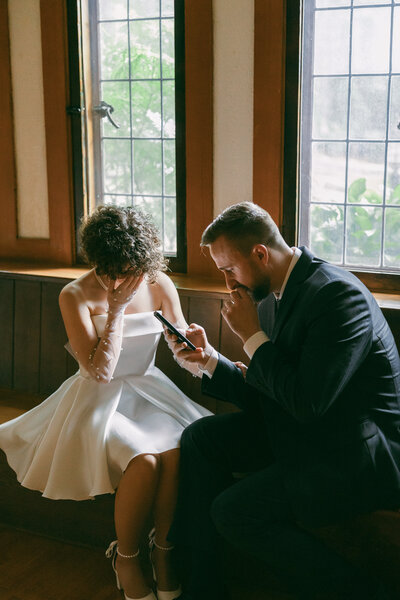 A woman in a short white wedding dress holds her face in her hand while a man in a blue suit reads from his phone with emotion