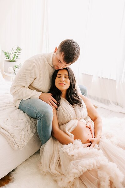 portrait of a husband and wife  for maternity photography. She is sitting on the floor leaning on his legs and he is leaning down and cuddling her wearing a white sweater.