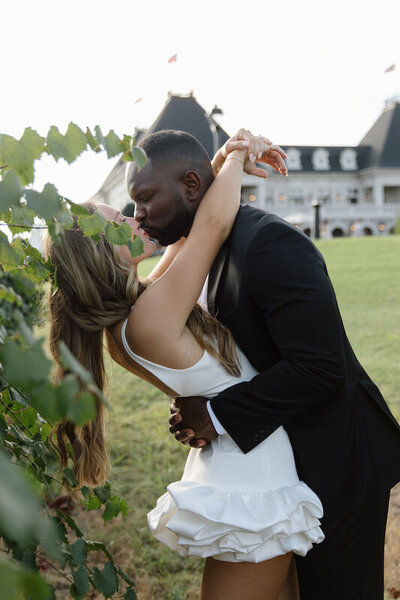 A couple kisses next to vines with a big mansion in the background