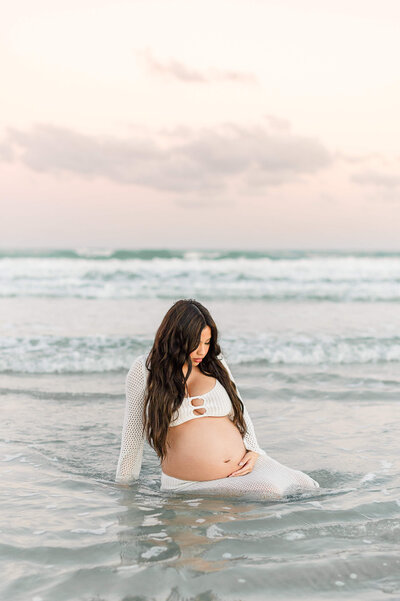 Pregnant mother sits in the water at sunset holding her belly during maternity photos