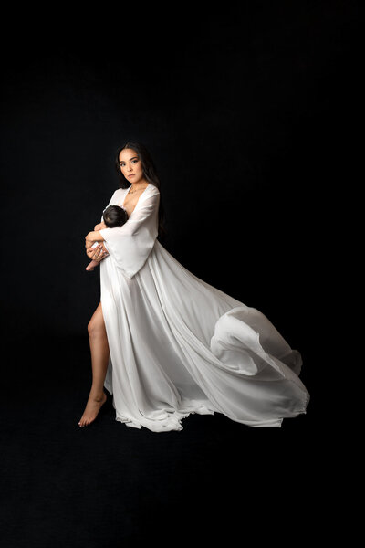 A mother is holding her newborn baby  while wear a flowing gown for a motherhood picture that is taken in a photography studio in Asheville North Carolina