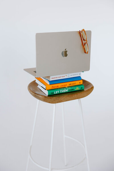 Laptop with eyeglasses hanging on it sitting on top of a stack of business coaching books on a stool