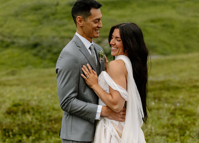 Bride and groom laughing together during their mountain elopement in Alaska, with lush green hills in the background.