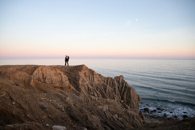 Newly engaged couple kiss on huge ocean cliff with ocean, setting sun and moon in the background in Montauk, NY