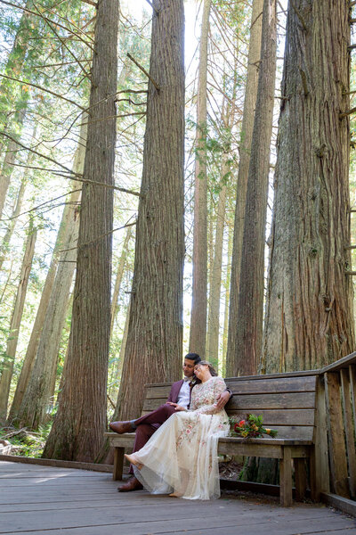 Bride rest her head on her husbands shoulder while they sit on a bench with towering trees all around.