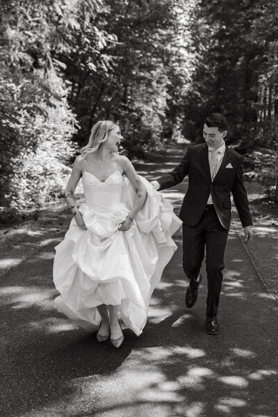 Groom holds his bride's train as they playfully run down a path among towering pines during their Cleveland, Ohio wedding. 