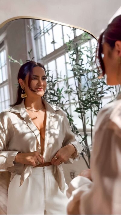 Emily Michele smiling while adjusting her beige outfit in front of a mirror — reflecting self-awareness, confidence, and authenticity through personal growth.