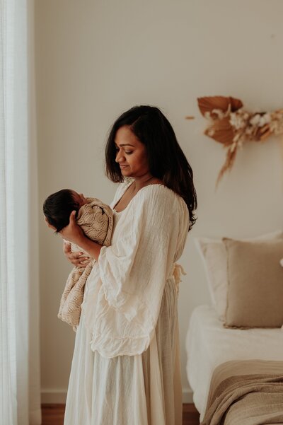 Woman with dark brown hair  holds her newborn baby and sweetly looks at him as she poses for a newborn session in a pretty studio
