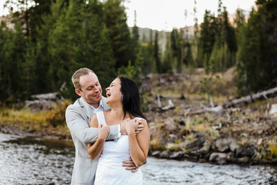 A blond groom wearing a gray jacket and white shirt wraps his arm across his Asian bride's chest as she grips his arm and looks back and him laughing and smiling while standing on the banks of the creek flowing near Sparks Lake in Bend, Oregon. | Erica Swantek Photography