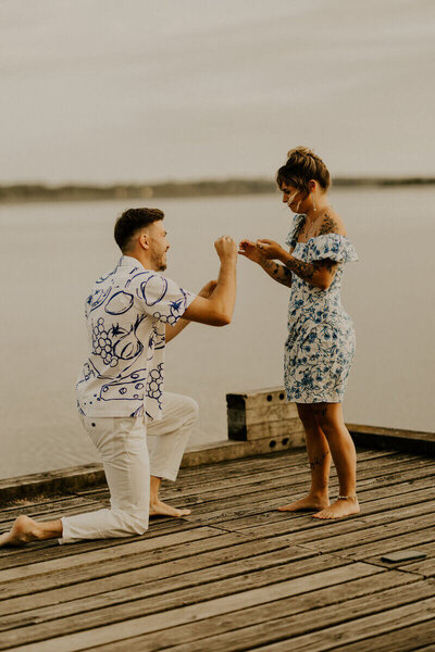 Clément, genoux à terre, fait sa demande en mariage à Laura sur un ponton en bois au bord du lac.