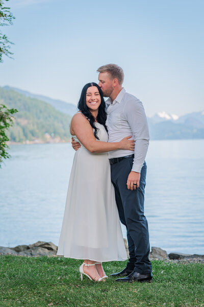 Groom kissing the brides forehead lake and mountains in the back round.