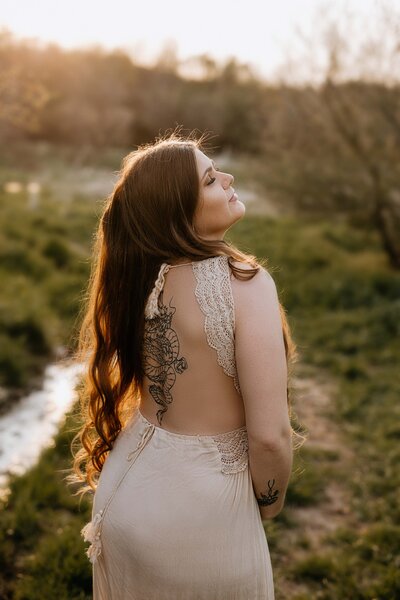 Expectant mom in London, Ontario field. Mom in open-back lace gown showing a tattoo on her back. She is looking over her shoulder with soft like creating a halo behind her.