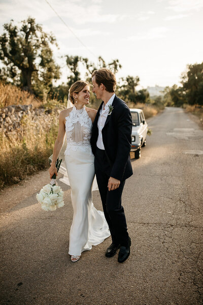 Bride and groom walk holding each other on the country road of Masseria Moroseta with a vintage Fiat  bridal car in 5he backdrop