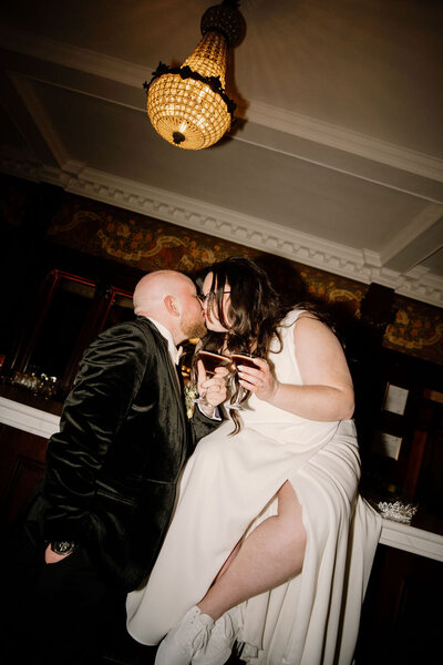 Newlywed couple sharing a kiss and holding champagne glasses during their indoor New York City wedding, photographed by Dom Augustine.