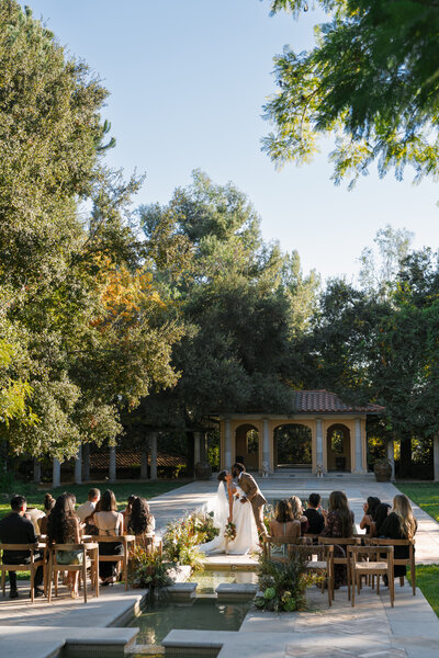 Bride and groom kissing in a lush garden setting as guests look on, set against a classic Mediterranean-style building, enhancing the romantic atmosphere of the wedding at Etienne Estate in Pasadena, Los Angeles.