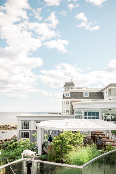 A view of the Cliff House Maine’s bright white exterior and waterfront pergola, with lush greenery and a sparkling ocean backdrop under a sunny sky.