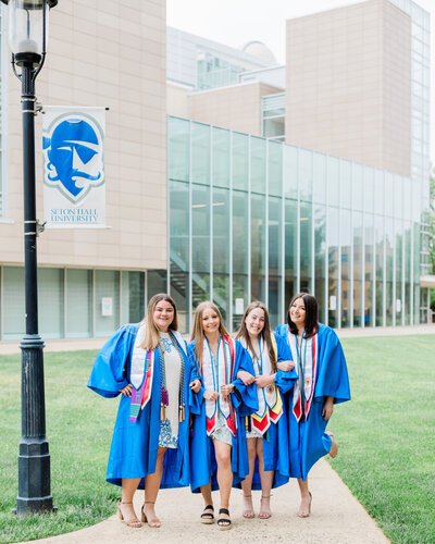 Four girls in blue graduation gowns posing in front of a Seton Hall University building