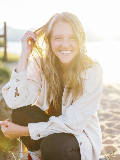 A woman smiling at the camera and kneeling on a beach during sunset with sun flare during her brand photoshoot