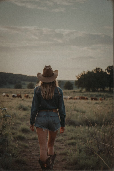 A person wearing a cowboy hat, denim shirt, and shorts walks through a grassy field toward a herd of cattle in the distance, embodying the authentic spirit that inspires Sweet Pea Ink Creative’s brand design approach.